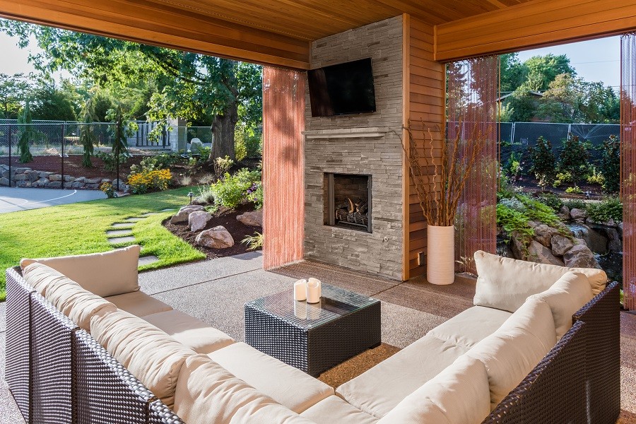 Brightly lit covered outdoor space with a comfortable couch facing TV and fireplace protected by outdoor automatic shades.  