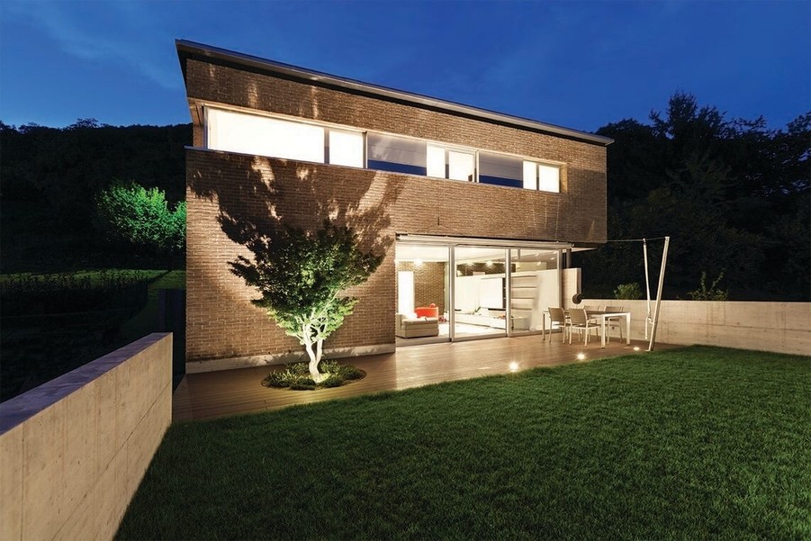 Modern two-story house with brick exterior, large glass sliding doors, and a lit backyard patio featuring a tree and outdoor dining area at dusk.