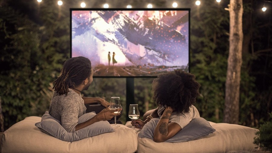 Two people relaxing in an outdoor home theater with glasses of wine and string lights above.
